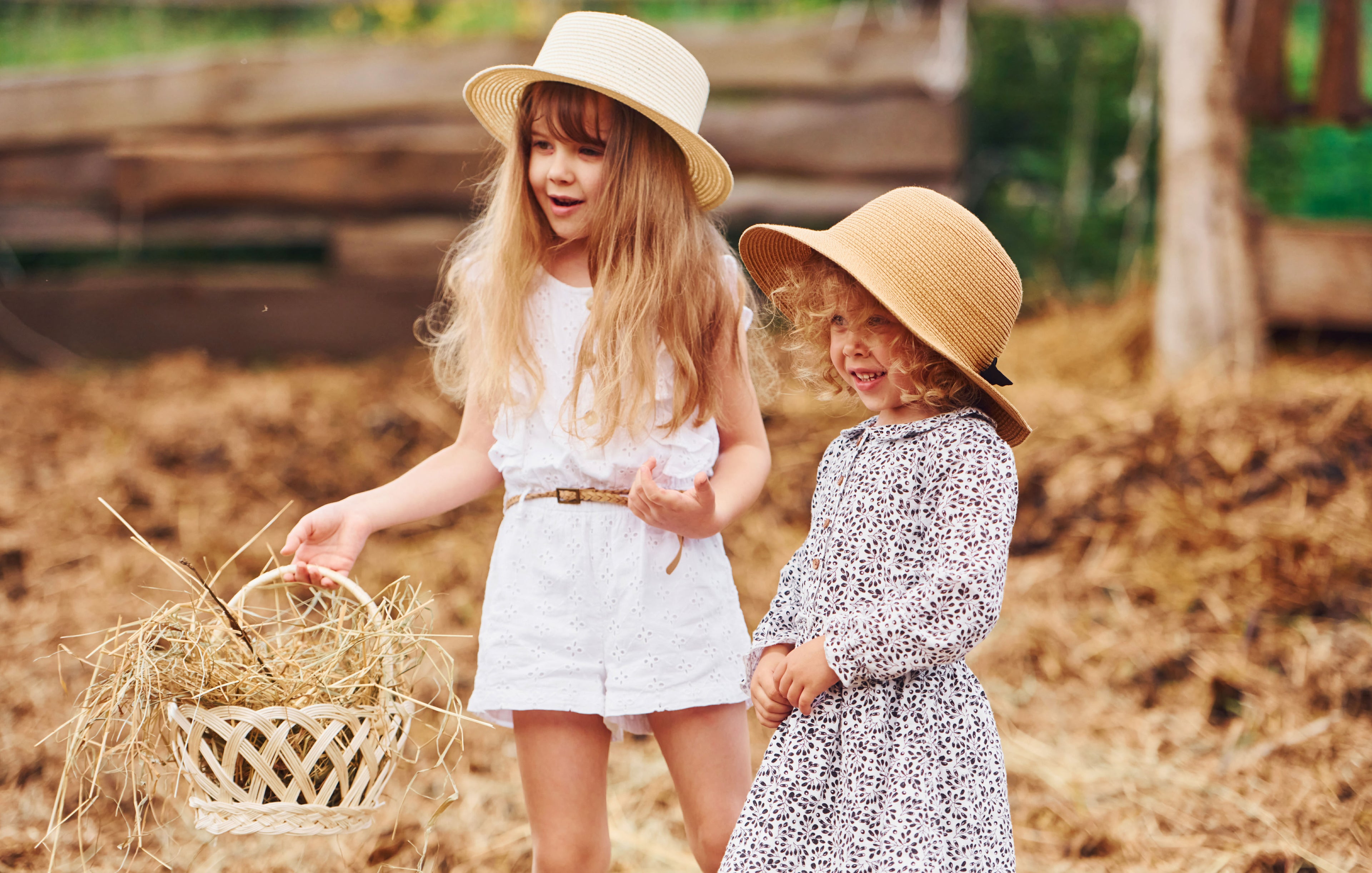 Two young girls in straw hats standing in a field with hay.
